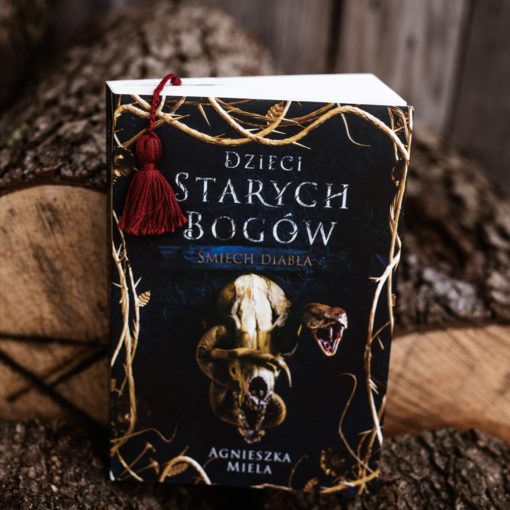 Na zdjęciu zdjęcie okładki książki Dzieci Starych Bogów. Śmiech diabła, w tle kawałki drewna z korą