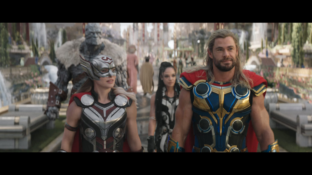 Fotos z filmu. Przedstawia on głównych bohaterów przechadzających się po mieście bogów. Na pierwszym planie znajdują się Thor oraz Jane Foster. Oboje są w swoich boskich zbrojach. Za nimi idą Walkiria oraz Korg.