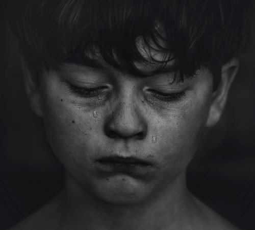 Na czarno białym zdjęciu chłopiec, po którego twarzy spływają łzy, usta ma zagryzione, wzrok spuszczony